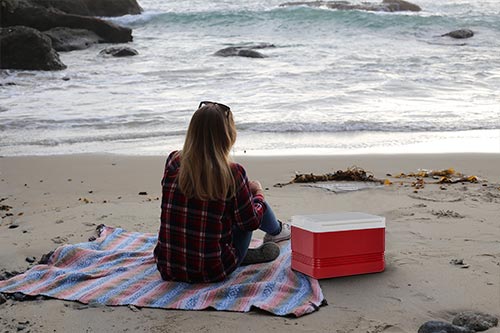 Chica frente al mar viendo las olas con toalla y nevera roja de playa pequeña